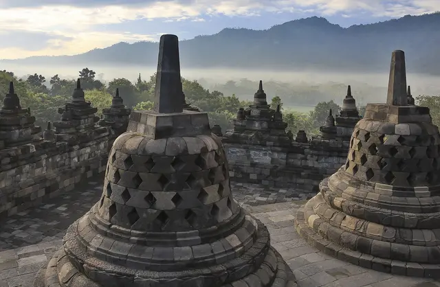 Latticed bell-shaped stone stupas on the upper terrace of Borobudur, with misty jungle and hills beyond