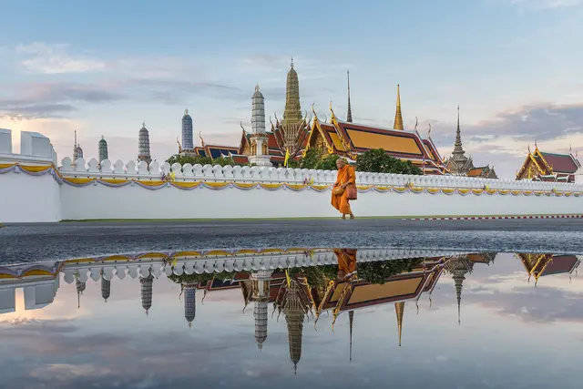 A monk in saffron robes walking past the white walls and golden spires of the Grand Palace in Bangkok, reflected in a puddle