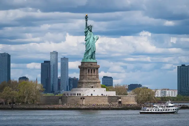 The Statue of Liberty on Liberty Island with skyscrapers behind and a sightseeing boat passing