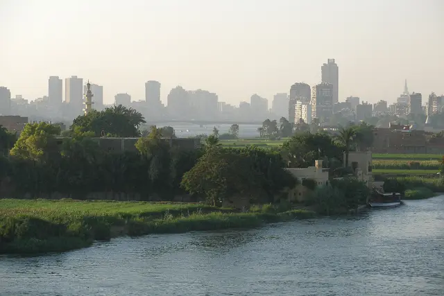 The Cairo skyline seen across the Nile, with palm trees and green riverbanks in the foreground