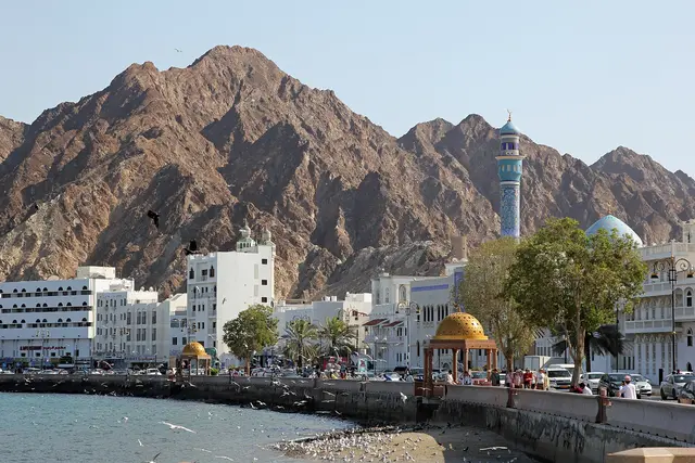 White buildings, a blue-tiled minaret and golden-domed pavilions along the Mutrah Corniche in Muscat, below rugged brown mountains